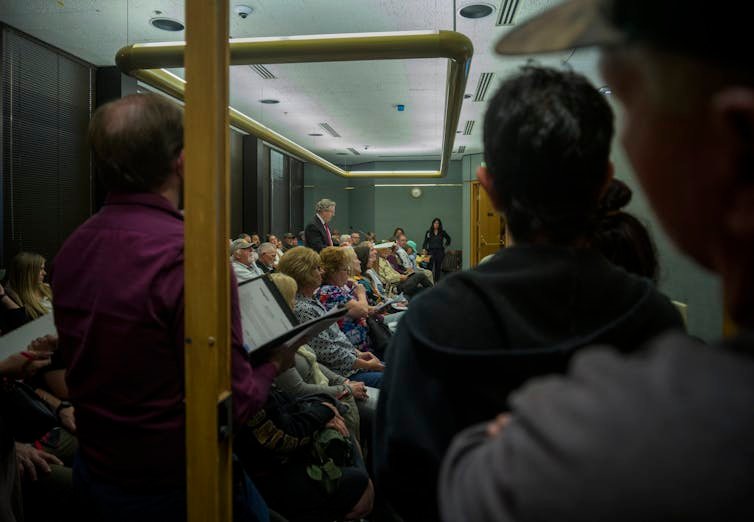 A crowd listens to a speaker in a public meeting room.