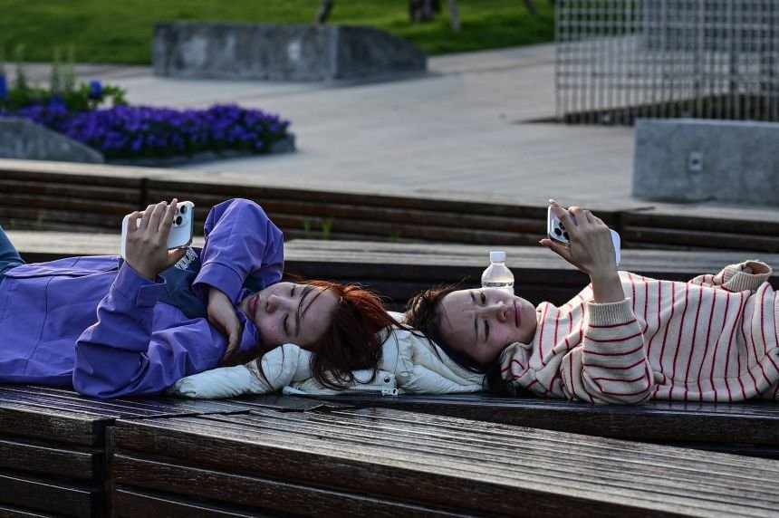 Two women lying on a bench look at their phones by the Huangpu River in Shanghai on April 9, 2026.