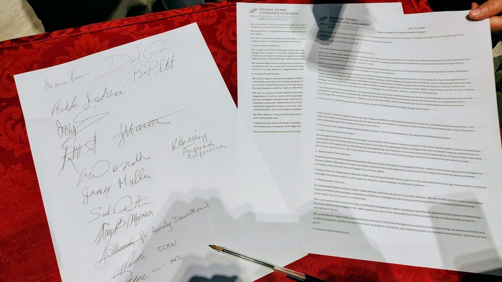 Two documents on a red table: one page with several signatures, one with densely printed text and a pen between them. A person's hand can be seen in the upper right corner.