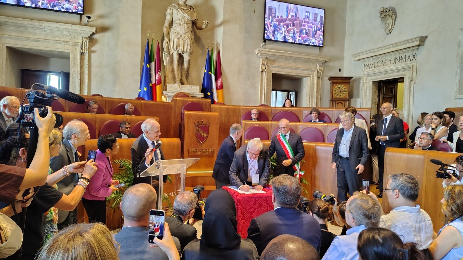 A man signs a document at a red-covered table in a crowded hall, in the presence of officials, photographers and onlookers.