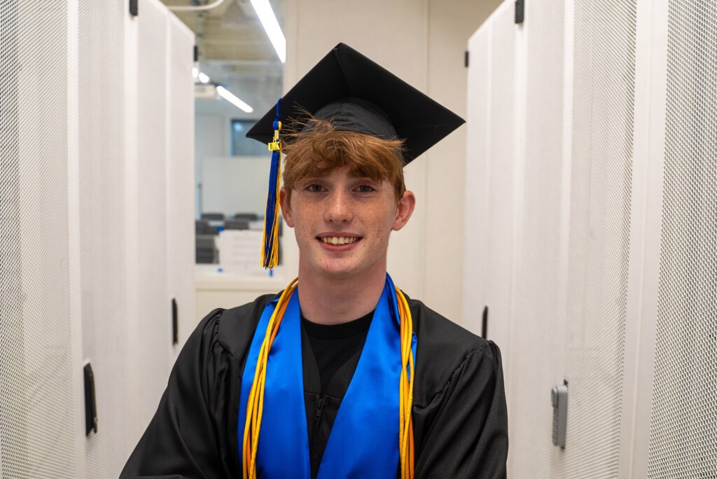 Thomas Safago poses for a photo in Discovery Hall wearing his graduation cap and gown.