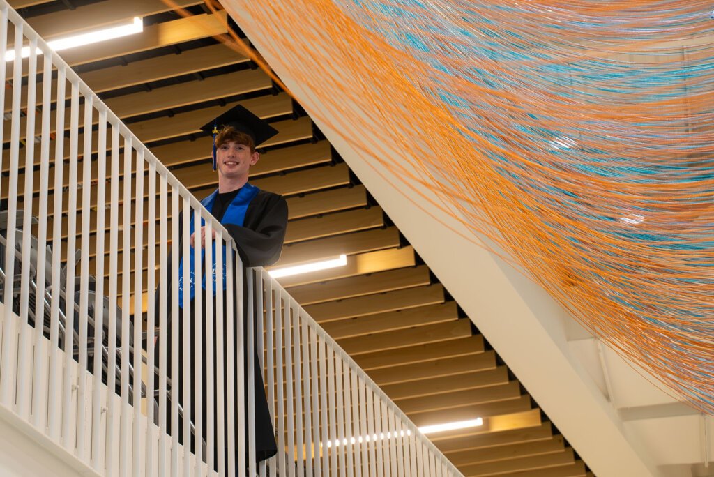 Thomas Safago poses for a photo in Discovery Hall wearing his graduation cap and gown.