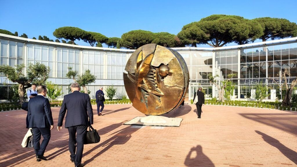Several people in business attire walk toward a large spherical bronze sculpture in a modern courtyard surrounded by glass buildings and trees.