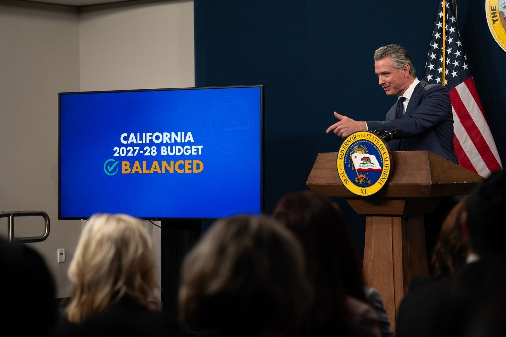 A suited official speaks at a podium during a press conference, framed by blurred figures in the foreground. An American flag and part of the California state seal appear behind them as they gesture with their left hand while addressing the audience. A monitor with the words 