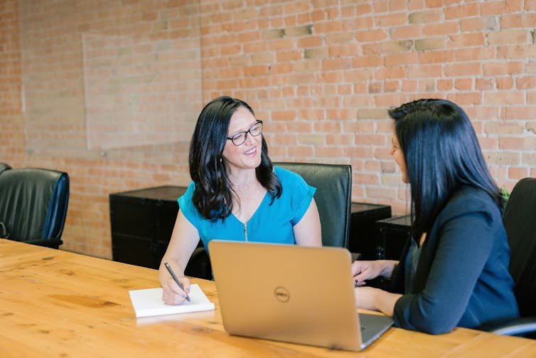 Two women are talking at a table, next to them is a laptop and a notepad.