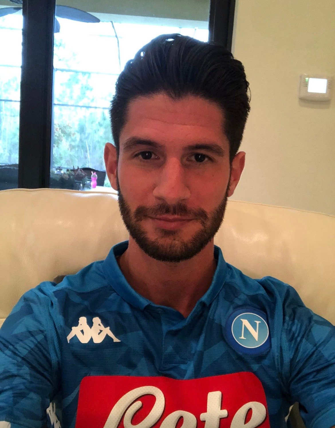 Meet Zachary Golba | Lead Software Engineer and Creator of Via – SHOUTOUT MIAMI 5 Young man with dark hair and beard, wearing a blue sports jersey, sitting on a beige couch indoors.