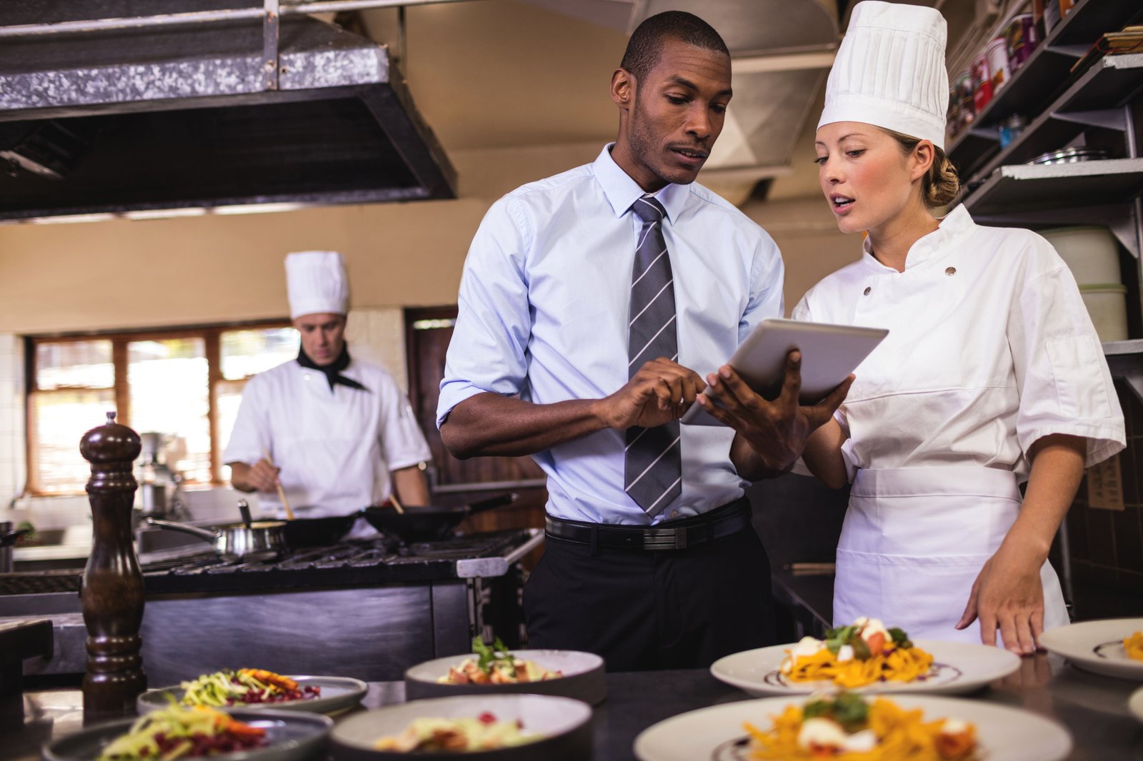 A chef and a manager talk using a tablet computer in the kitchen of a restaurant.