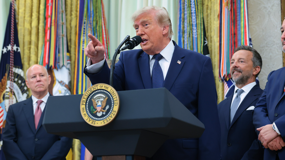 Donald Trump, a white man in a blue suit, points as he gives a speech from behind the podium while other men stand behind him.